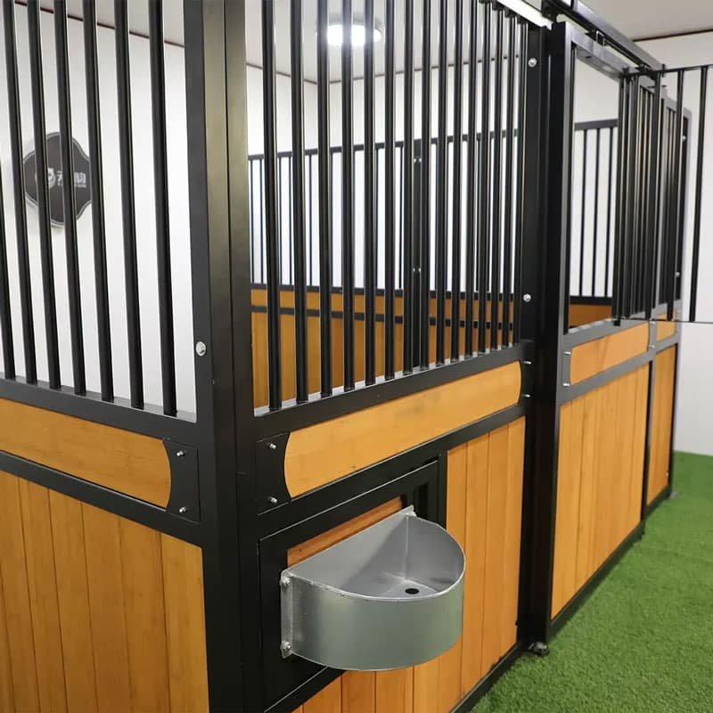 A series of horse stalls lined up within a stable. The stalls feature a combination of black metal bars and wooden panels, with overhead lights illuminating the space.