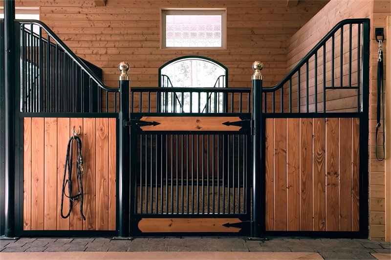 An interior view of a horse stable with multiple stalls, each labeled with names. The stalls feature wooden doors with metal bars, and the bright interior is complemented by decorative lighting.