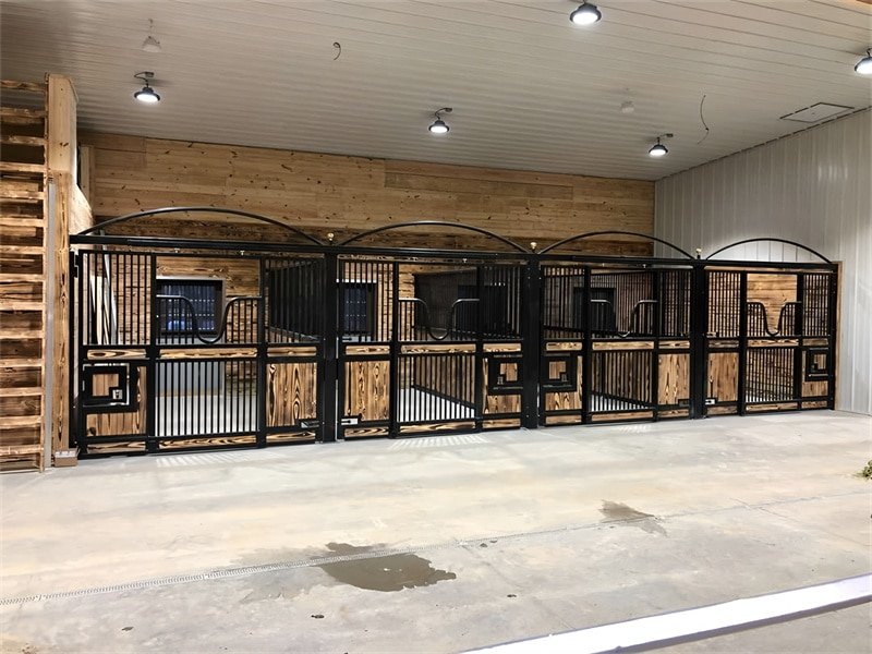 An interior view of a horse stable with multiple stalls, showcasing a combination of wood and metal materials, well-lit and organized.