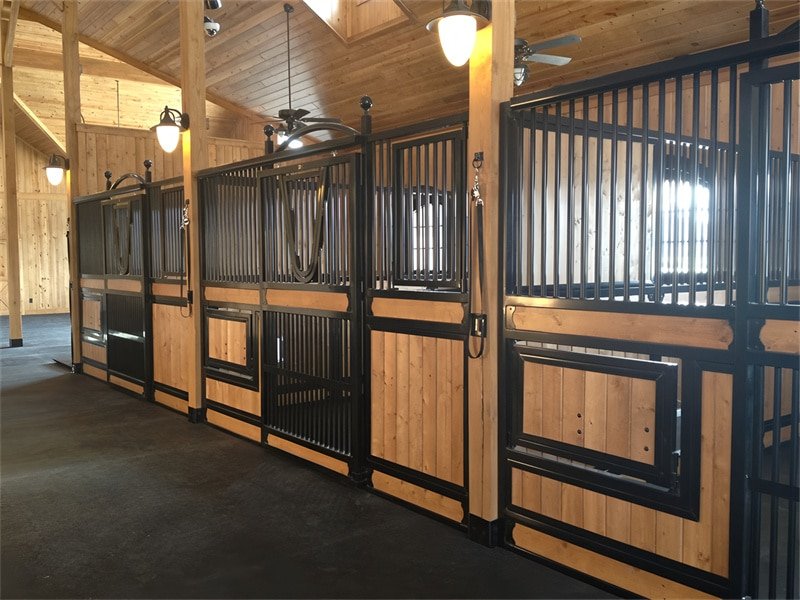 A series of horse stalls lined up within a stable. The stalls feature a combination of black metal bars and wooden panels, with overhead lights illuminating the space.