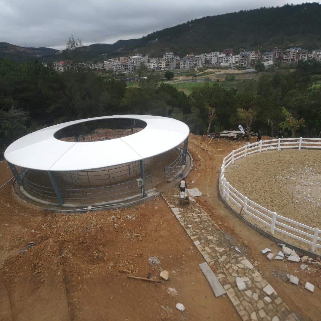 Aerial view of a circular horse walker with a white ring roof and metal rail enclosure beside a sand round pen at a hillside site under construction